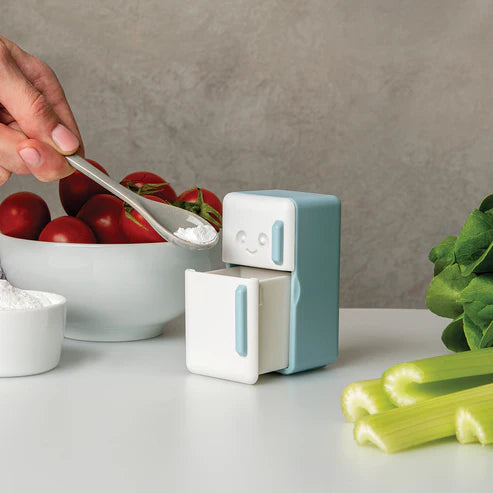 Small robot-like container with a face on a kitchen counter next to tomatoes and celery.