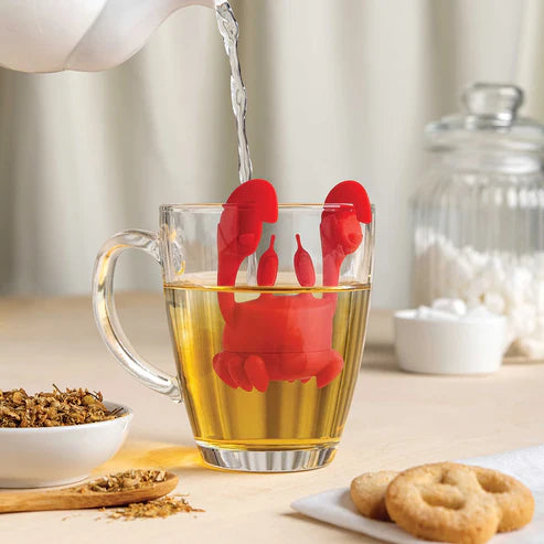 Red lobster-shaped tea infuser in a glass of tea with water being poured, on a table with tea leaves and cookies.